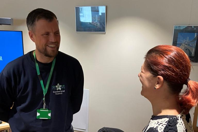 A man wearing a Cumberland Council lanyard talks to a lady in an office.